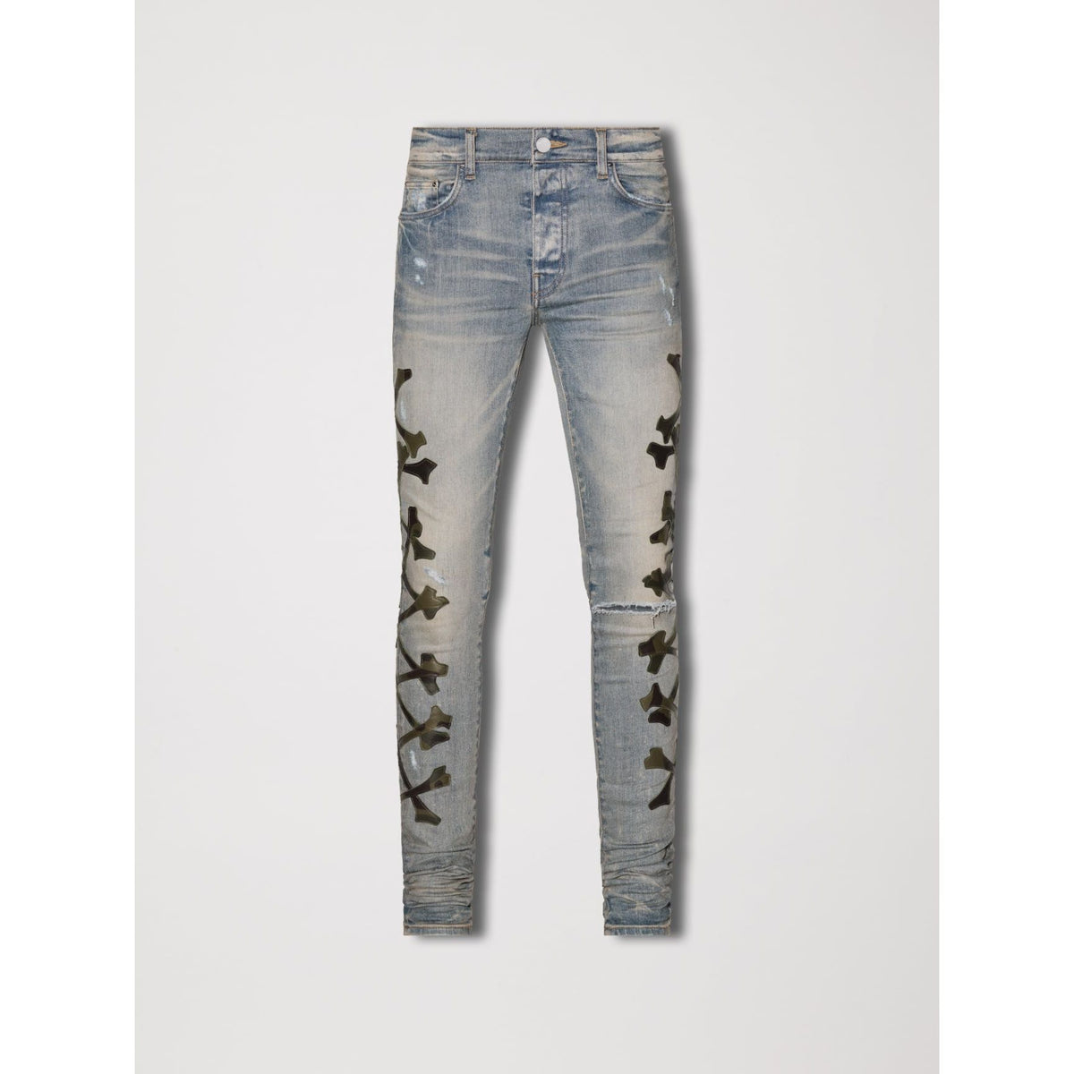 AMIRI Clay Indigo skinny jeans, distressed with a ripped right knee, and vertical dark green camo bone patches on both legs.