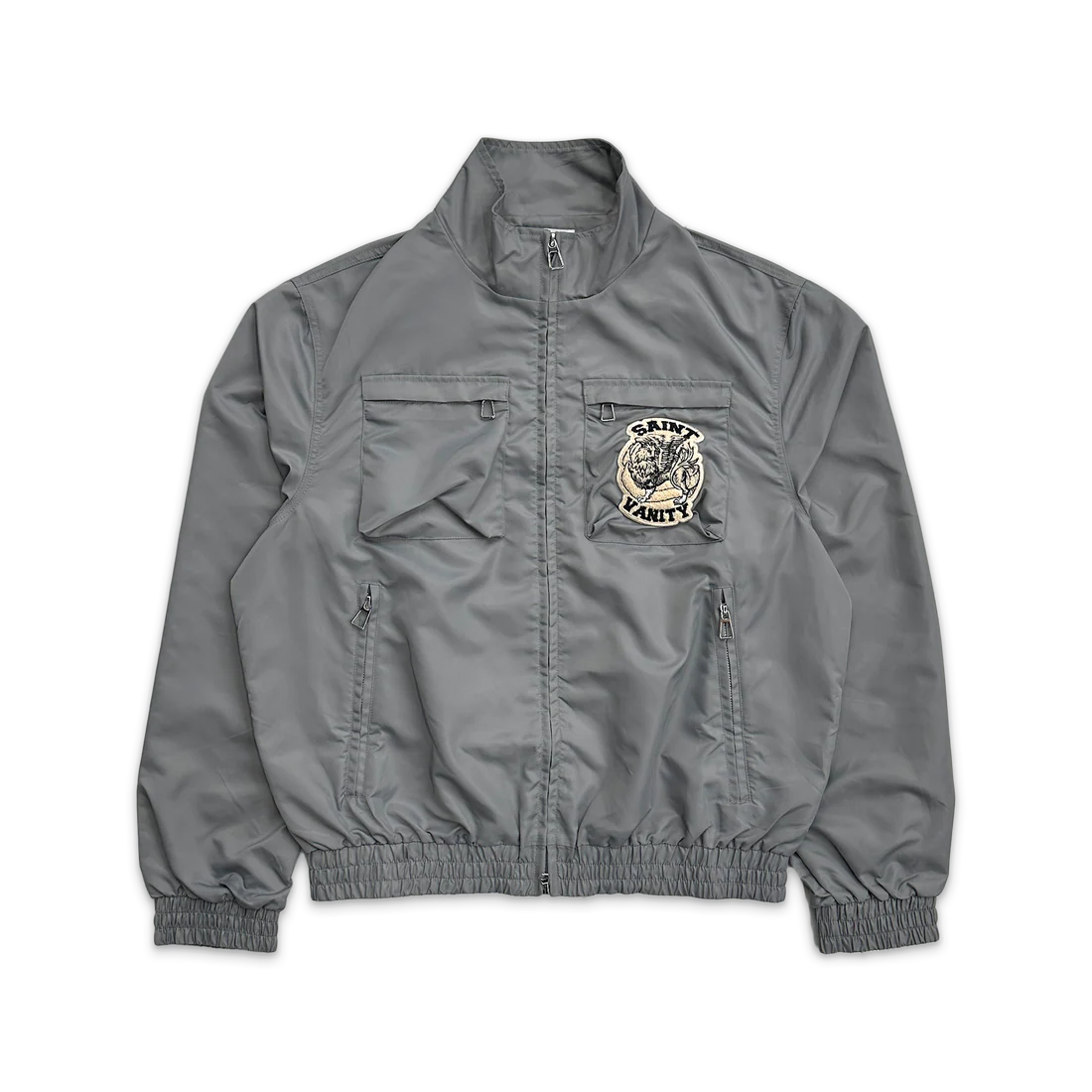 Grey nylon zip-up jacket with stand collar, ribbed cuffs/hem. Features chest flap pockets, lower zip pockets, & a Saint Vanity logo patch.
